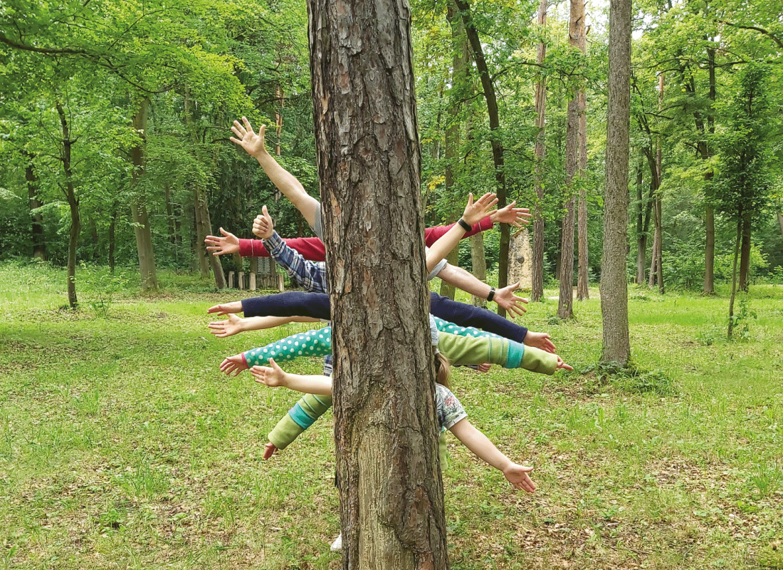 Kinder stehen mit ausgebreiteten Armen hinter einem Baumstamm mitten in einem lichten Wald